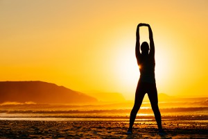 Fitness woman exercising and stretching arms on beautiful summer sunset or morning at beach. Female doing relaxing yoga and breathing exercises.
